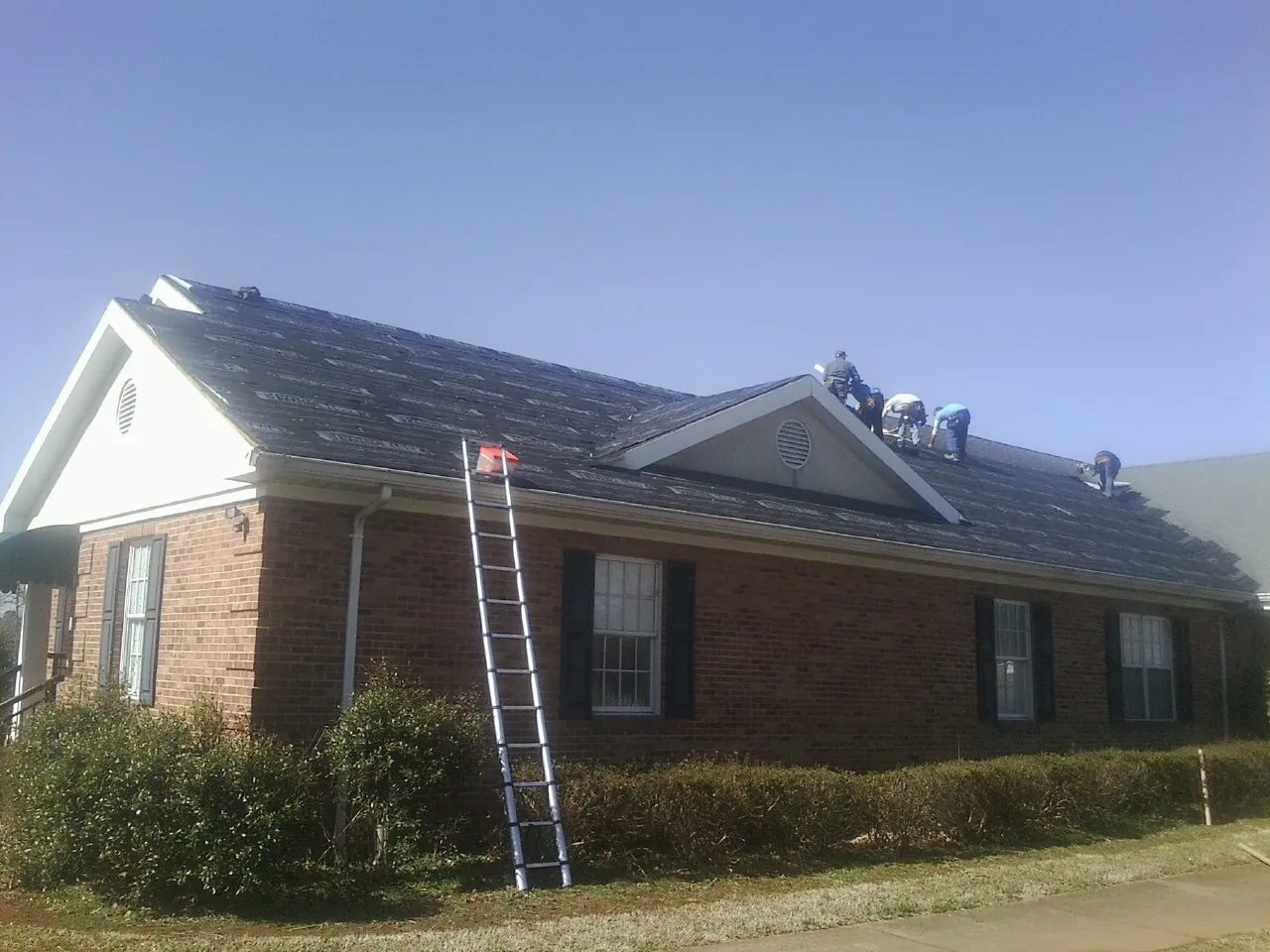 Crew tearing shingles off a brick ranch roof with a ladder set — Metal Roof Repair work by our crew in Clendenin