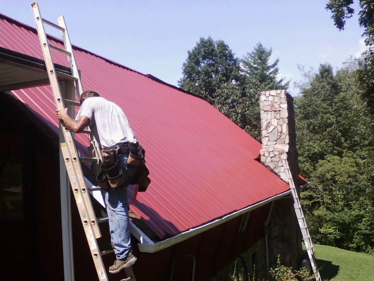 Roofer on a ladder beside a red metal roof and stone chimney — Soffit & Fascia Repair by Roofing Brothers in Clendenin